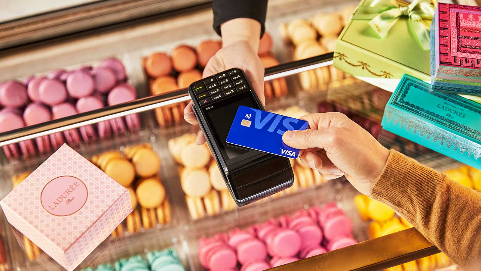 A display of cookies is visible in the background as a person holds up a Visa contactless payment card for payment. 